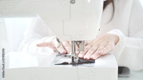 Female hands sew on a white sewing machine close-up. Concept of sewing in modern bright studio, woman in white sweater sewing grey cloth
