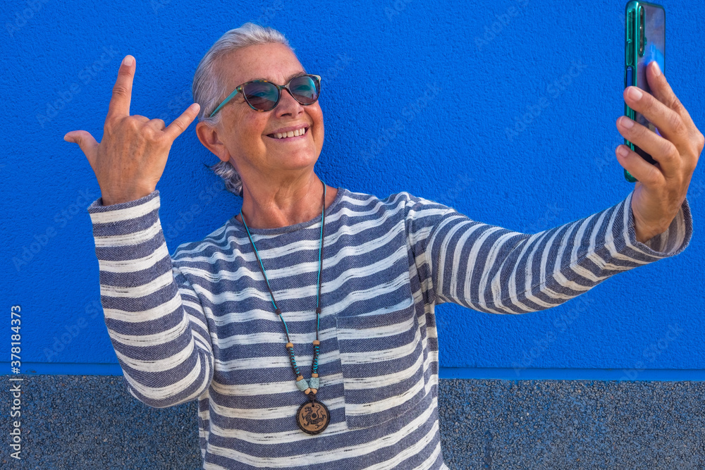 Naklejka premium Cheerful attractive senior woman with white hair smiling happy making a selfie using smart phone standing - blue background - tech and social elderly people