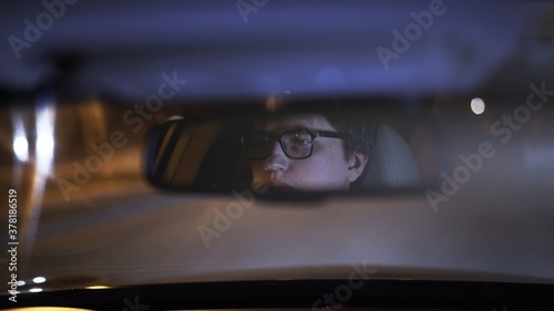Driver's face is reflected in a rear view mirror, night shot, violet color. Tired concentrated driver in eyeglasses is reflected in a mirror in car, night road shot driving all night