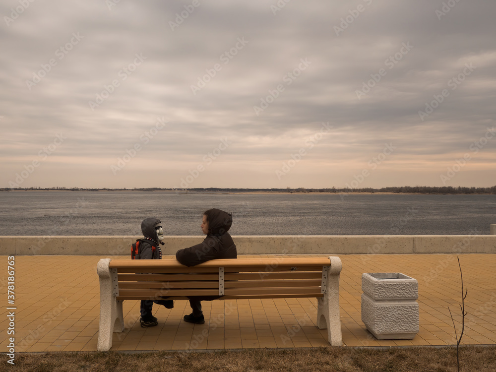 Adult woman with child in scary mask sits on wooden bench by river and looks at cloudy sky