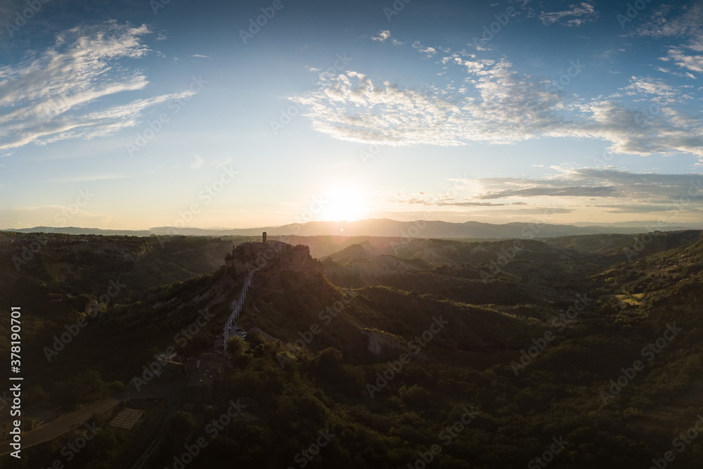 Fototapeta premium Aerial view of stone town Civita di Bagnoregio with the sun at the sunrise with clay badlands and trees in background