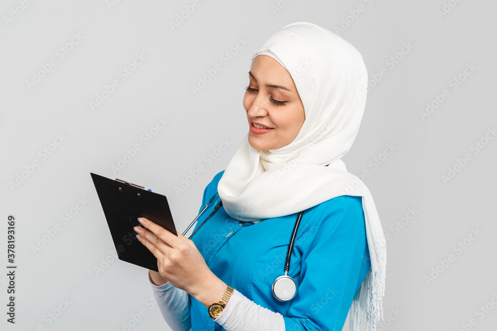 friendly, Muslim doctor or nurse woman in a hijab with a stethoscope writes  a prescription on pills on a gray background. Stock-Foto | Adobe Stock