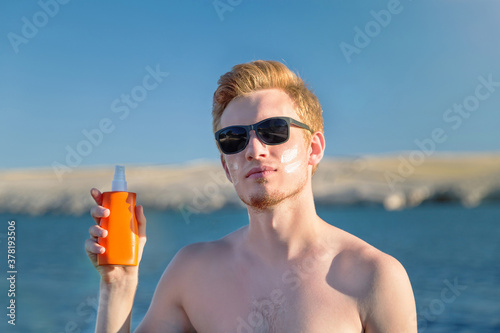 Attractive man with botle of protective sun cream (solar cream, sunscreen ). Young handsome caucasian male model with red hair on the natural background.