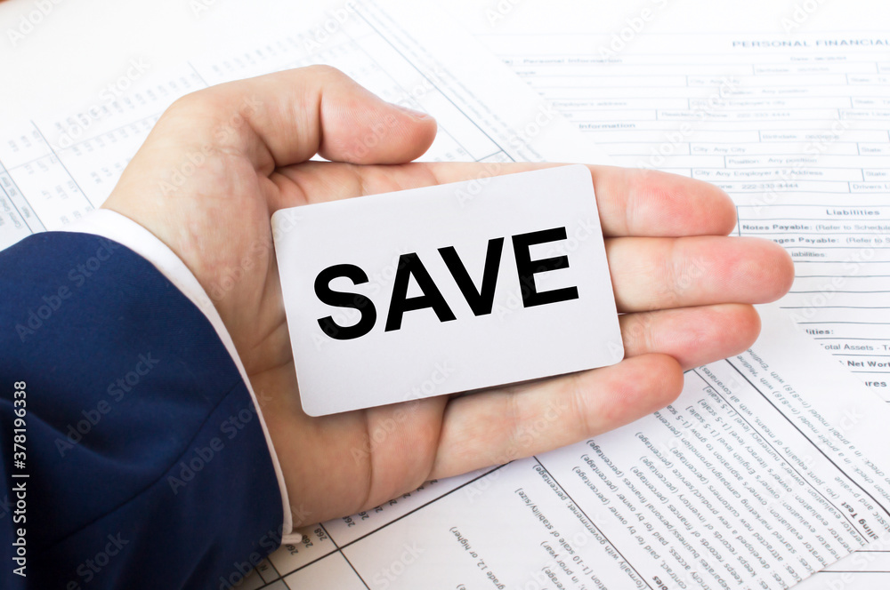 A man's hand is on the financial tables holding a business card with the inscription Save A man's hand is on the financial tables holding a business card with the inscription Save
