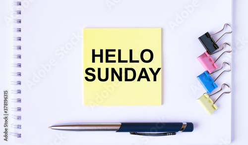 Yellow sticker with text Hello Momday lying on a white Notepad with a pen and paper clips
