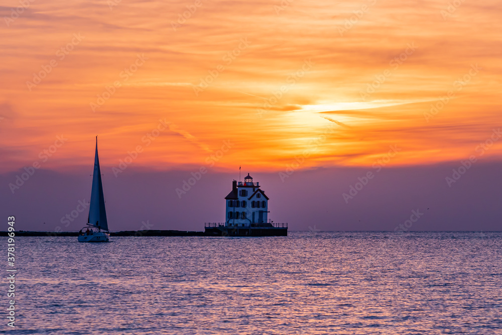 Sunset over calm water with sailboat sailing past lighthouse