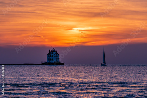 Sunset over calm water with sailboat sailing past lighthouse