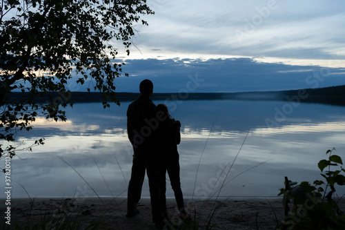 A couple in love - a man and a woman standing on the shore, in a romantic setting, admire the reflection of the evening clouds in the water surface of a forest lake.