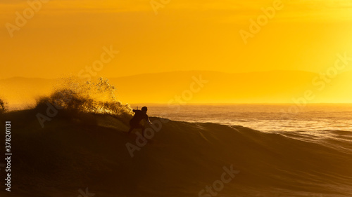 silhouette of a surfer riding a wave at sunrise with orange glow in background © Louis