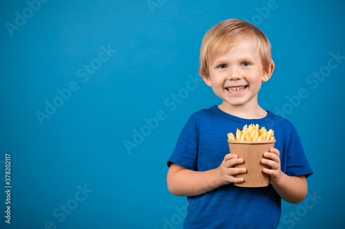 Boy blond child 4 years old happy holding a cup with fries in his hands. Close-up studio shot on blue background with space for text.