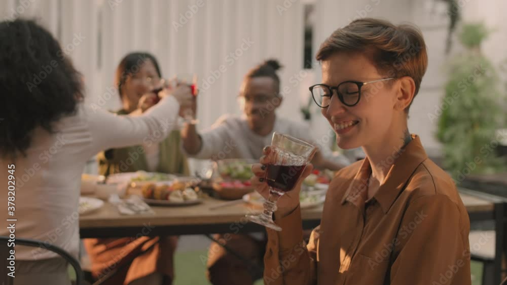 Medium shot of young Caucasian woman with short hair is sitting near dining table with wine glass in her hands, looking at camera while her friends clinking glasses on background Medium shot of young Caucasian woman with short hair is sitting near dining table with wine glass in her hands, looking at camera while her friends clinking glasses on background