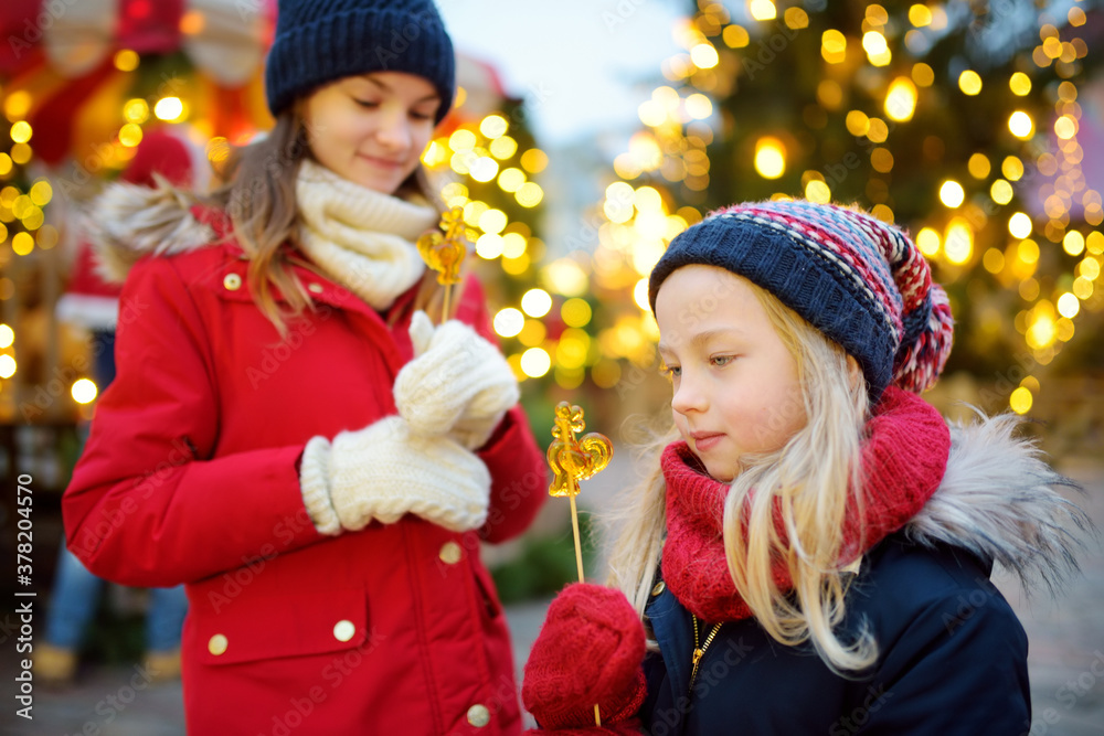 Two adorable sisters having rooster-shaped lollipops on traditional Christmas fair in Riga, Latvia. Children enjoying sweets, candies and gingerbread on Xmas market. Two adorable sisters having rooster-shaped lollipops on traditional Christmas fair in Riga, Latvia. Children enjoying sweets, candies and gingerbread on Xmas market.