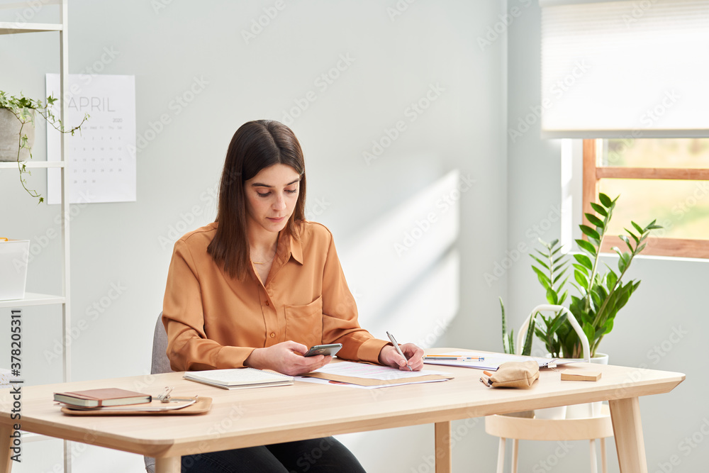 Young businesswoman browsing smartphone while making notes at home Young businesswoman browsing smartphone while making notes at home