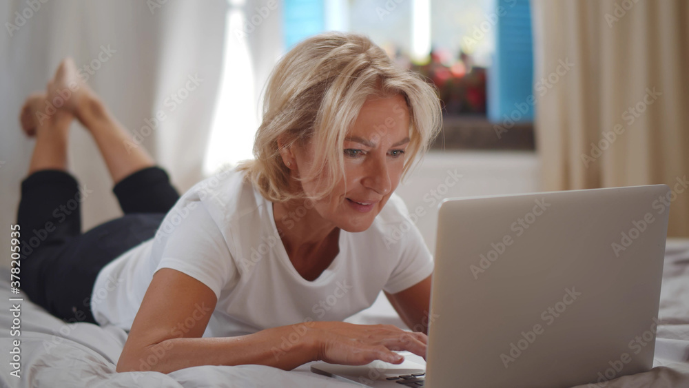Senior beautiful woman chatting on laptop lying on bed in morning Senior beautiful woman chatting on laptop lying on bed in morning