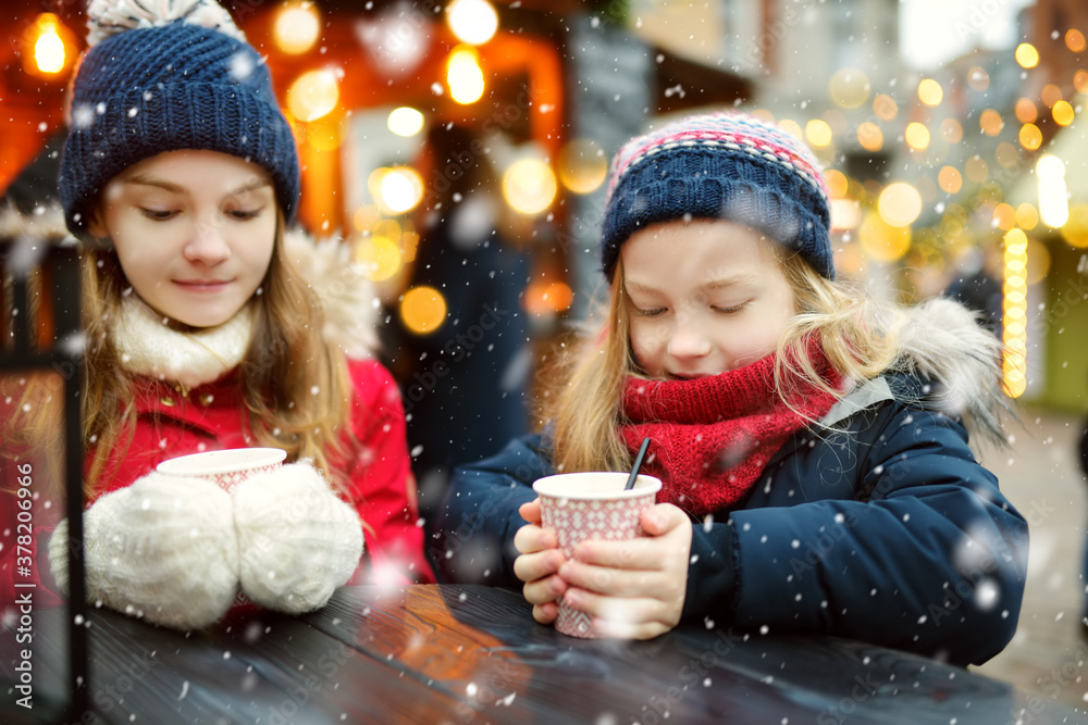Two adorable sisters drinking hot chocolate on traditional Christmas fair in Riga, Latvia. Children enjoying sweets, candies and gingerbread on Xmas market. Two adorable sisters drinking hot chocolate on traditional Christmas fair in Riga, Latvia. Children enjoying sweets, candies and gingerbread on Xmas market.