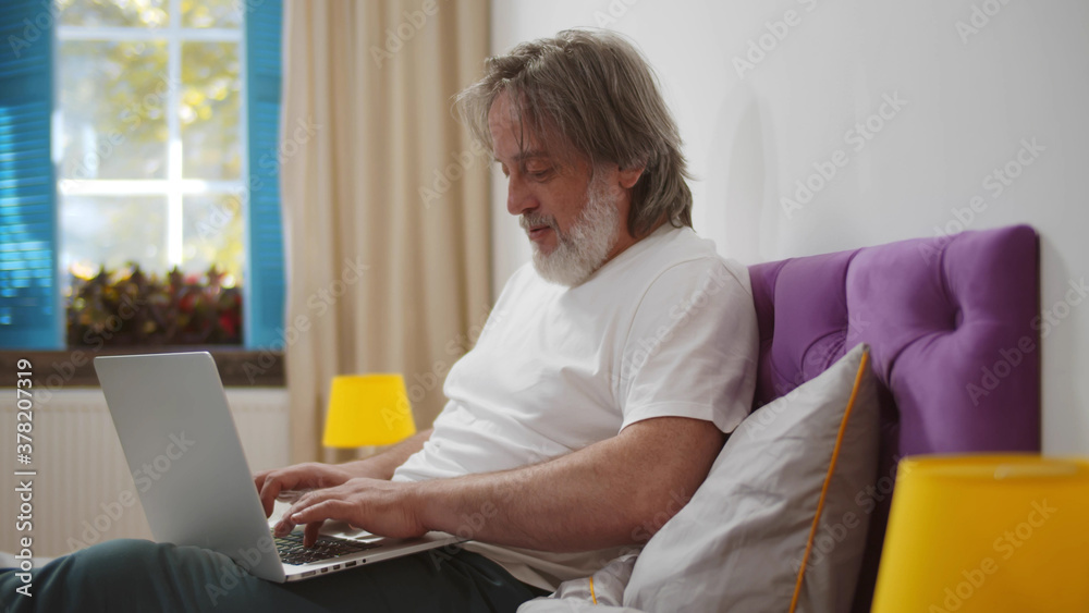 Senior man sitting on bed working online from home with laptop Senior man sitting on bed working online from home with laptop