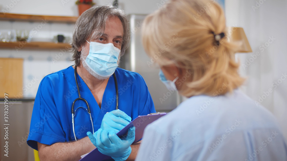 Doctor in mask writing prescription for senior female patient during home consultation Doctor in mask writing prescription for senior female patient during home consultation