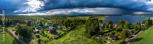 Aerial Panorama of Scandinavian pine tree forest landscape, summer village with Sunny and rainy areas, clear blue sky, white clouds, green yellow fields. View on typical landscape of Northern Sweden
