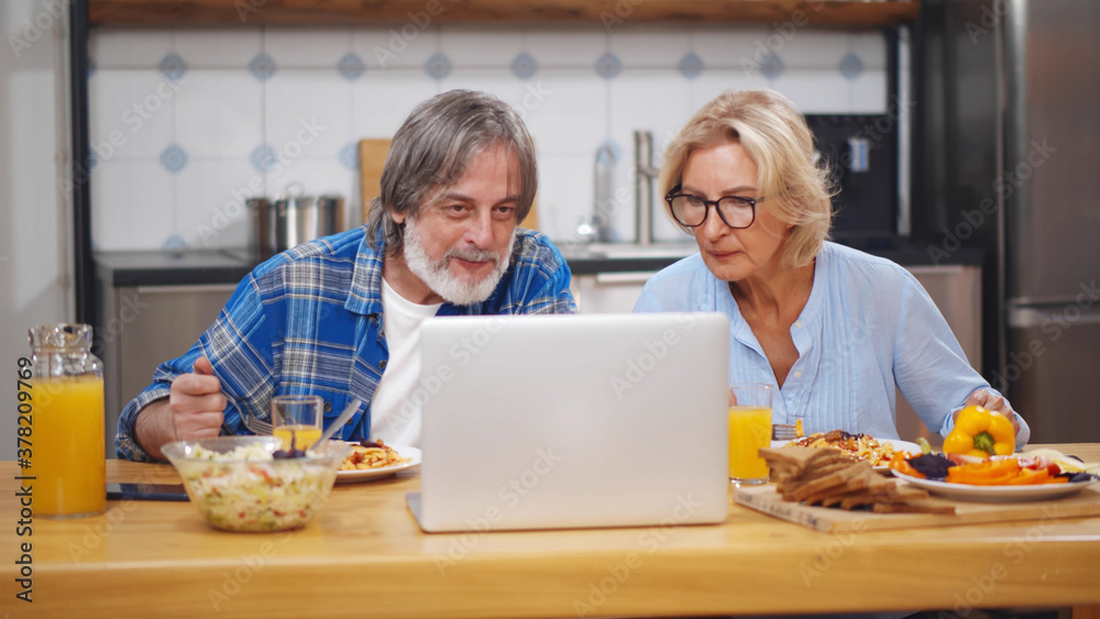 Happy mature loving couple sitting at kitchen and eating using laptop
