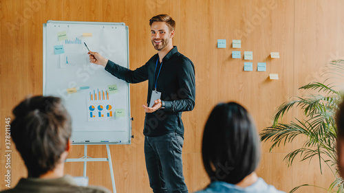Caucasian businessman giving presentation to colleagues pointing on flip chart while they sitting around watching him