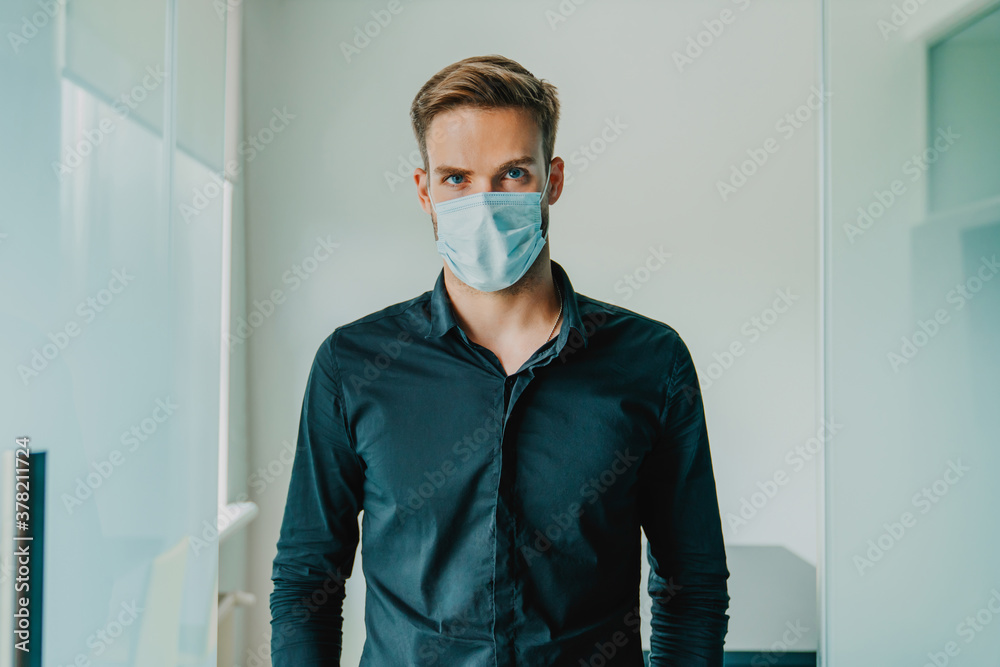 Portrait of young business man wearing medical protective mask and looking at camera standing in modern office. Working safely during pandemia Portrait of young business man wearing medical protective mask and looking at camera standing in modern office. Working safely during pandemia