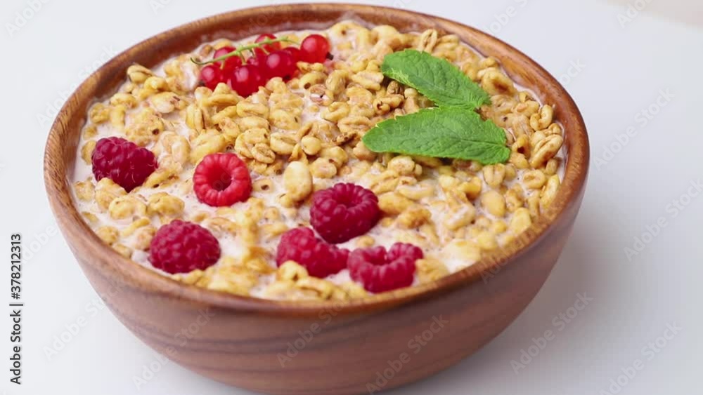 Rotating of wheat flakes porridge with milk, raspberry and currant in wooden bowl on white wooden background and linen textile. Side view, close up. Rotating of wheat flakes porridge with milk, raspberry and currant in wooden bowl on white wooden background and linen textile. Side view, close up.