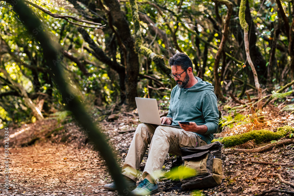 Adult man work with computer laptop and roaming phone connection in the middle of the green wild forest - concept of digital nomad and modern smart working people everywhere internet connected Adult man work with computer laptop and roaming phone connection in the middle of the green wild forest - concept of digital nomad and modern smart working people everywhere internet connected