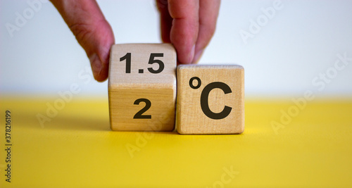 Symbol for limiting global warming. Male hand turnee a cube and changes the expression '2 C' to '1.5 C', or vice versa. Concept. Beautiful yellow table, white background, copy space.