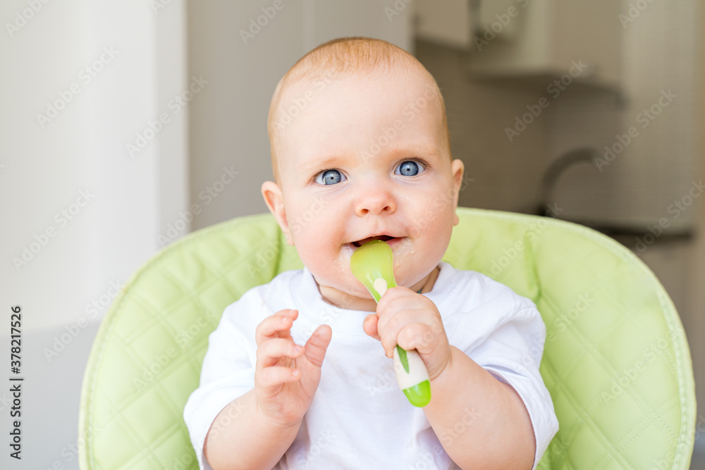 a small child with blue eyes and blond hair sits in a high chair and eats the first bait. Healthy diet. Baby 6 months a small child with blue eyes and blond hair sits in a high chair and eats the first bait. Healthy diet. Baby 6 months