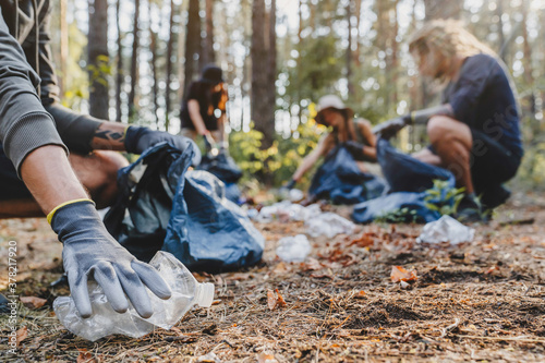 Close up of hands with plastic bottle with group of young people friends volunteers collecting plastic bottles to trash bags in forest background. Ecology concept photo