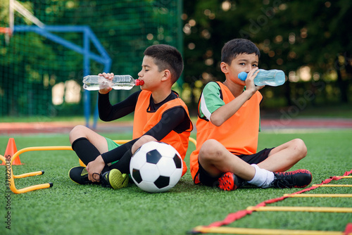 Two teen boys in football uniforms having a rest on sport field with artificial covering and drinking fresh water. photo