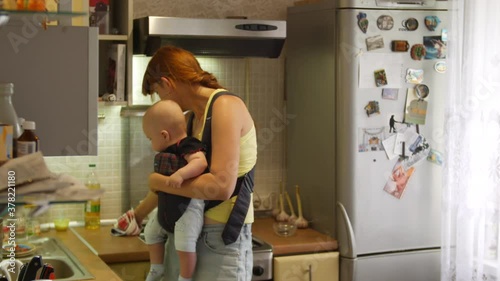 Woman with an infant boy sitting in a sling cooks soup for a dinner and cleans up at the kitchen and then kisses the kid. Mother household chores concept. Slow motion front view in 4k video.