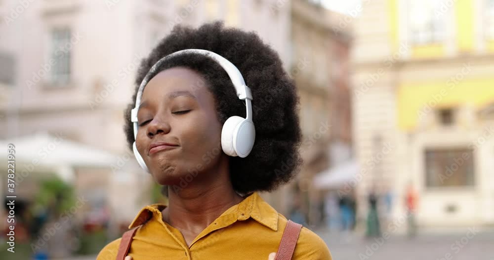 Caucasian happy beautiful African American female in headphones walking on street and listening to music outdoors. Joyful girl in good mood enjoying songs in town. Close up portrait. Leisure concept