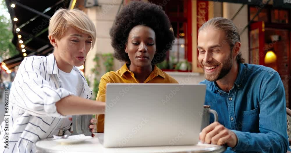 Portrait of cheerful mixed-races friends sitting at cafeteria terrace with coffee and talking while looking at laptop. African American girl and Caucasian man smiling. Blond female tapping on computer Portrait of cheerful mixed-races friends sitting at cafeteria terrace with coffee and talking while looking at laptop. African American girl and Caucasian man smiling. Blond female tapping on computer