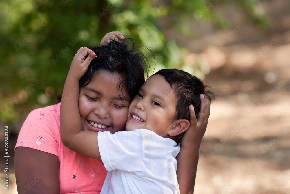 Little brother is hugging his older sister with his arms around her face and both are smiling in outdoor setting. Little brother is hugging his older sister with his arms around her face and both are smiling in outdoor setting.