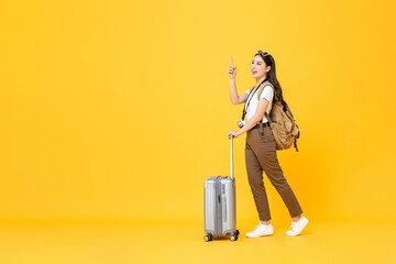 Full length travel concept portrait of beautiful smiling young Asian woman tourist with trolley bag pointing up isolated on colorful yellow studio background