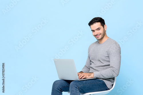Smiling handsome man sitting typing on laptop computer in isolated studio light blue background