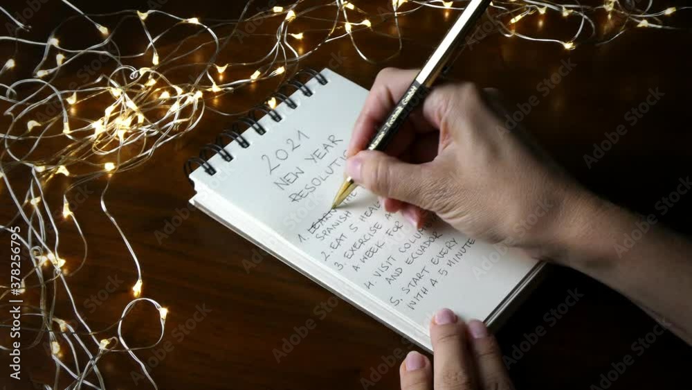 Female hands crossing out items from a list of specific detailed resolutions for the new year 2021, in a notebook in a festive setting with led lights on a dark wooden table.