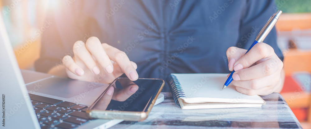 Woman is writing on a notebook with a pen and she is using a mobile phone in the office.