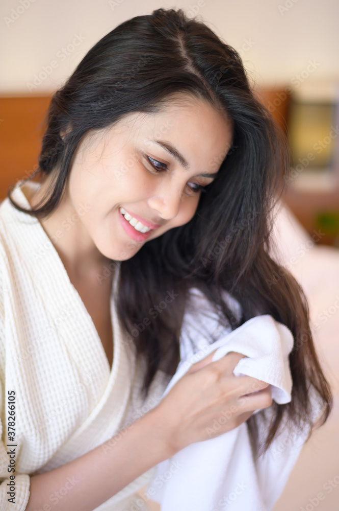 Smiling young woman wearing white bathrobe wiping her hair with towel after a shower in bedroom Smiling young woman wearing white bathrobe wiping her hair with towel after a shower in bedroom