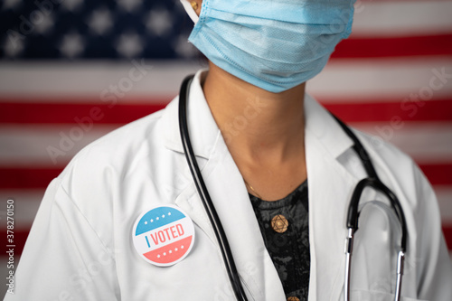 Doctor or nurse medical mask and stethoscope wearing placing I voted sticker to her apron - concept of US election with US flag as background. photo