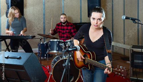 Attractive positive female soloist playing guitar and singing with her music band in sound studio