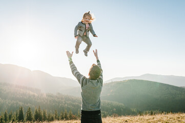Portrait of a father holds, throws up happy daughter on hands walking on nature in autumn day. Dad and child playing in the mountains. Concept of family spending time together on vacation.