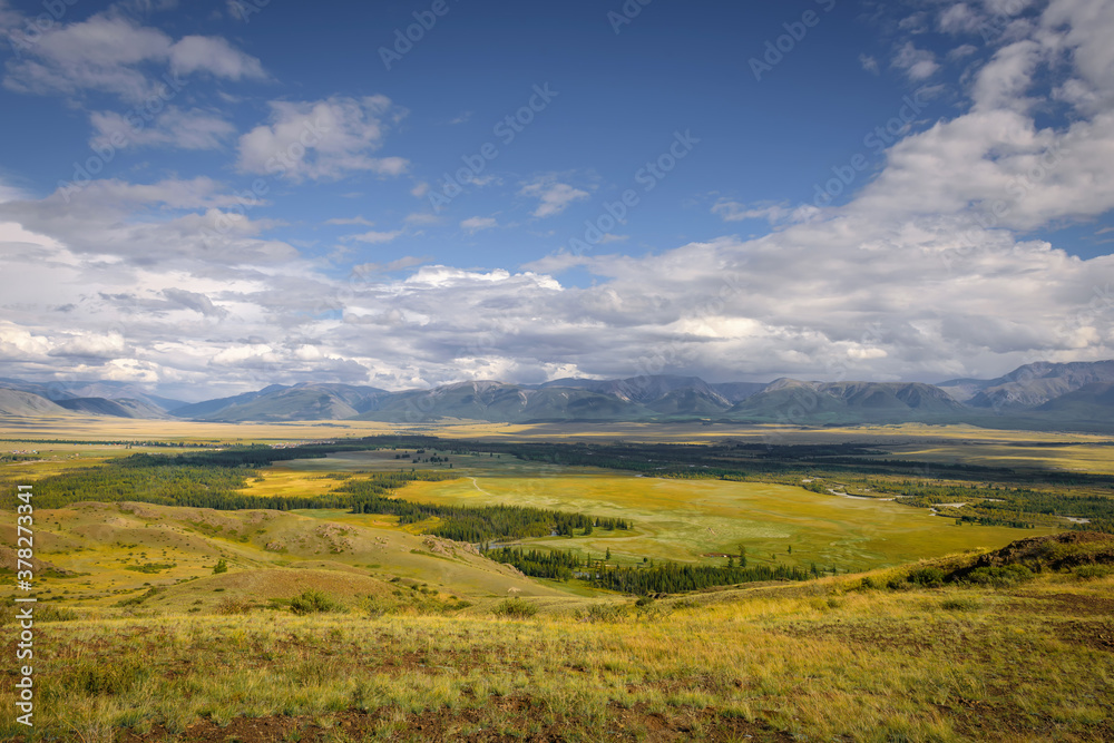 Picturesque green valley in sunlight against snowy mountain range under cloudy sky on summer or autumn day. Beautiful panorama of the Altai mountains, Siberia.