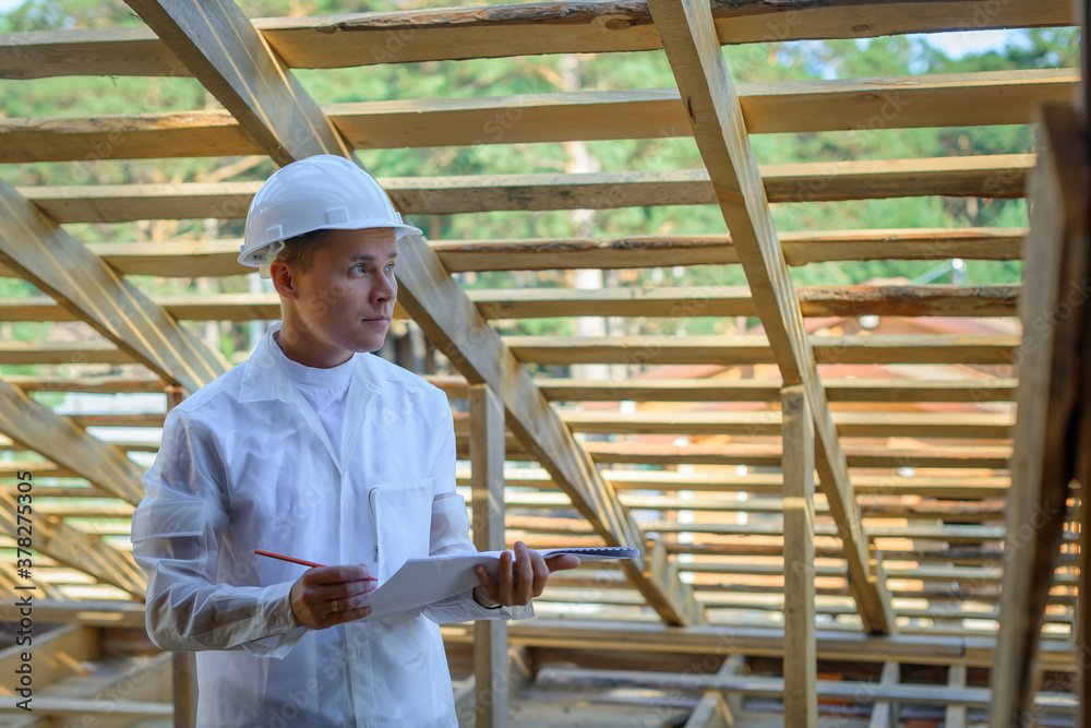 Attractive builder in the white helmet looks at the drawings. Foreman with a construction plan and a pen on the background of a cottage under construction. Portrait of architect with copy space.