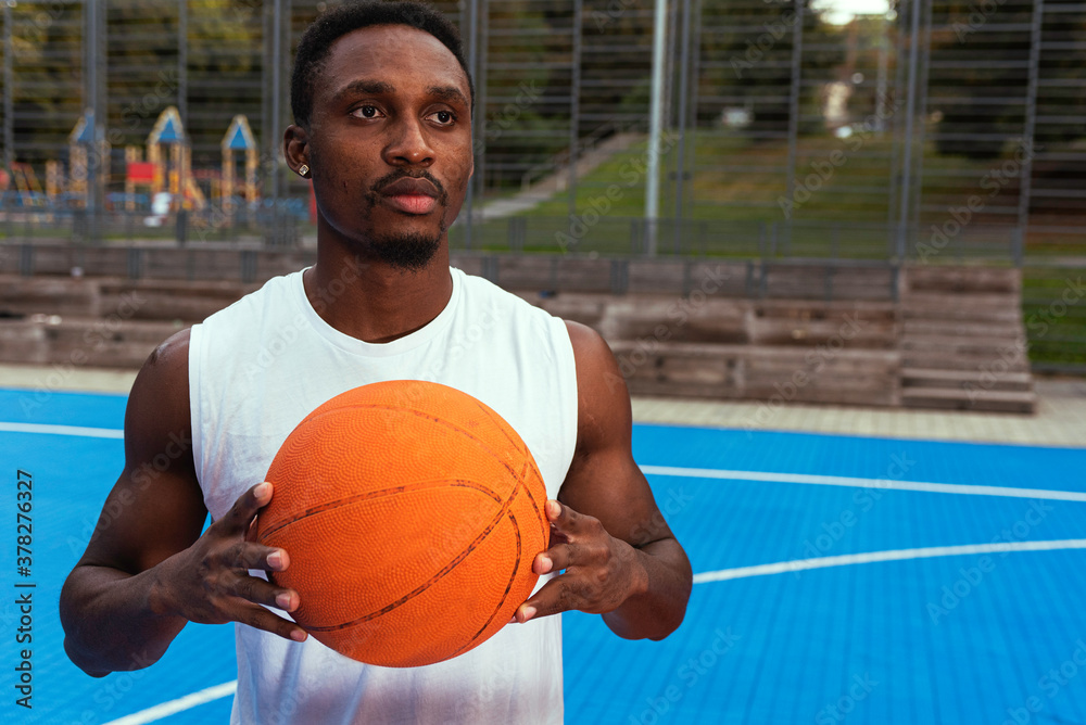 A man in a white T-shirt holds a basketball in his hands in front of him, looking straight at the playground. The concept of controversy, healthy living A man in a white T-shirt holds a basketball in his hands in front of him, looking straight at the playground. The concept of controversy, healthy living
