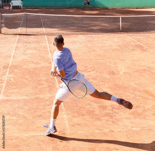Young tennis player on clay tennis court with racket plays forehand hit. Professional tennis player in dynamic motion after hitting the ball. Sports action frame. Back view, shadow, square size