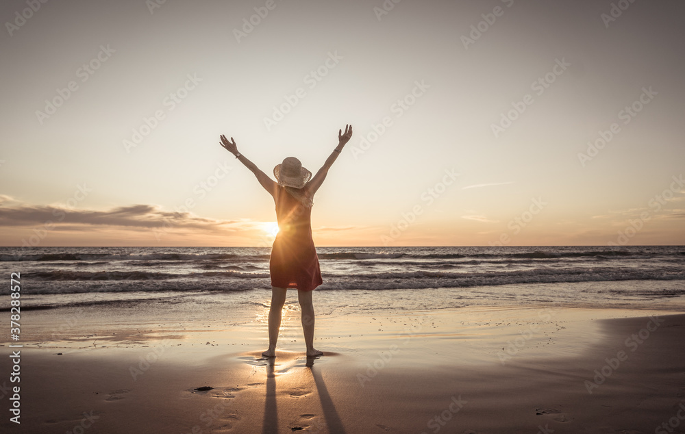 Woman in red with arms outstretched by the sea at sunrise enjoying freedom and life - obrazy, fototapety, plakaty 
