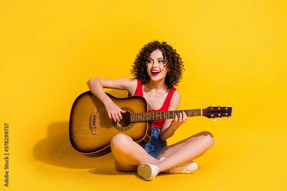 Portrait photo of hipster female musician with curly hair singing song holding keeping playing melody guitar chords smiling sitting down isolated on vivid yellow color background - obrazy, fototapety, plakaty 