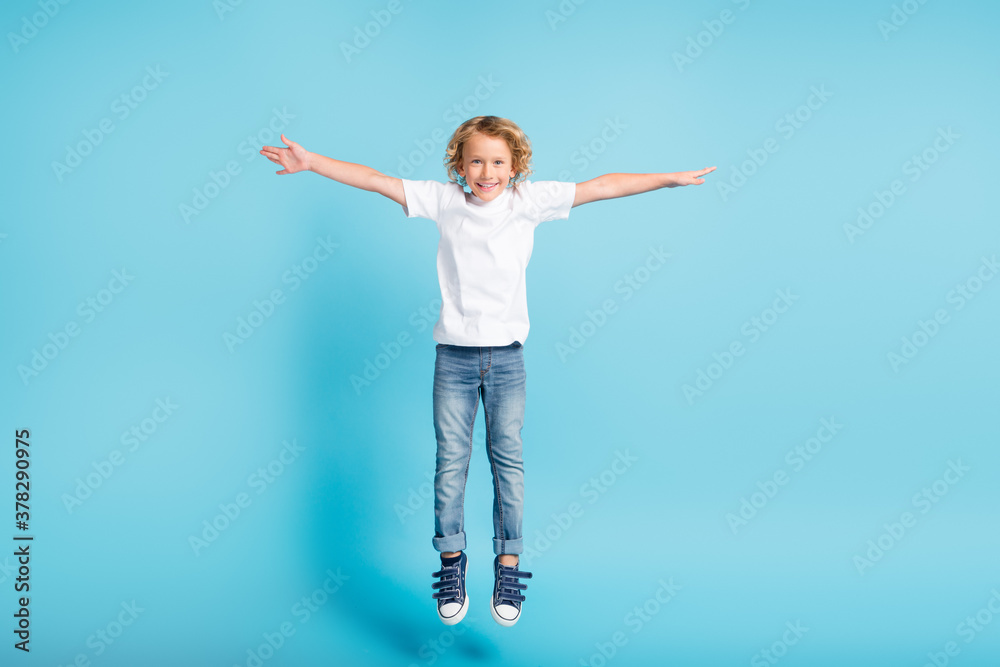 Photo of little boy jump raise hands wings wear white shirt jeans sneakers isolated blue color background Photo of little boy jump raise hands wings wear white shirt jeans sneakers isolated blue color background